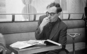 Black and white photo of Jean Luc Godard in a suit wearing glasses at a table