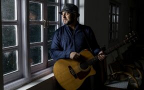 Singer-songwriter Rohan Solomon holding an acoustic guitar and staring out a window