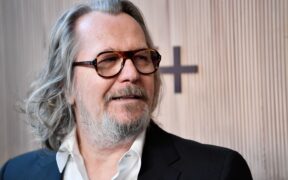 Gary Oldman in glasses and a blue blazer, with long grey hair