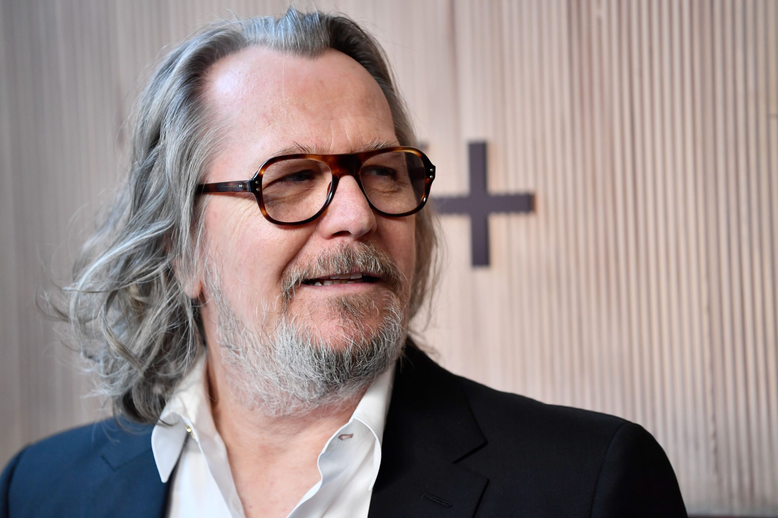 Gary Oldman in glasses and a blue blazer, with long grey hair