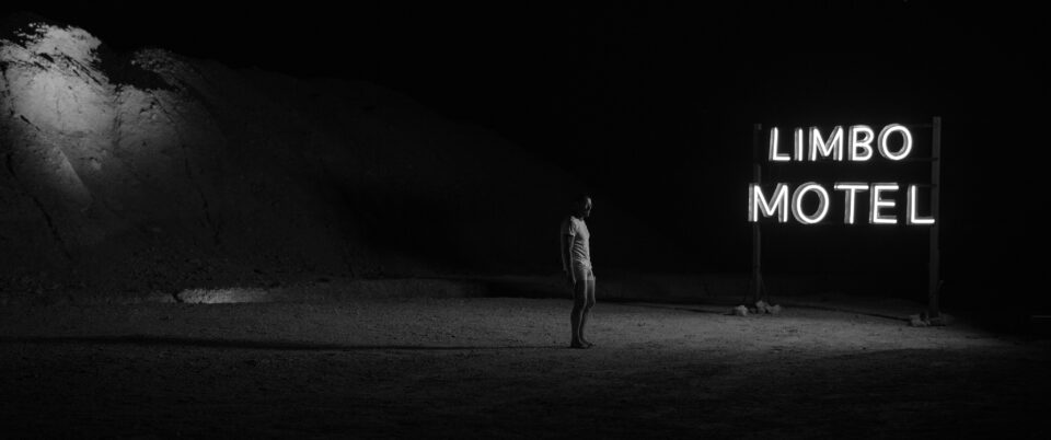 A dark black still from Australian movie 'Limbo' with actor Simon Baker in the center, in front of a neon sign 'Limbo Motel'