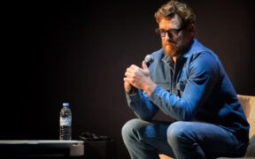 Actor Simon Baker seated in a blue long sleeve shirt and trousers, wearing glasses and holding a mic