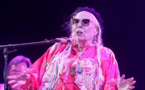 Joni Mitchell singing in dark glasses wearing red top