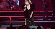 Kelly Clarkson singing into a mic on stage wearing a green ensemble with black jacket