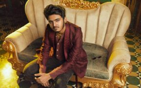 Sai Abhyankkar in red jacket sitting on a gold, white sofa for Katchi Sera video