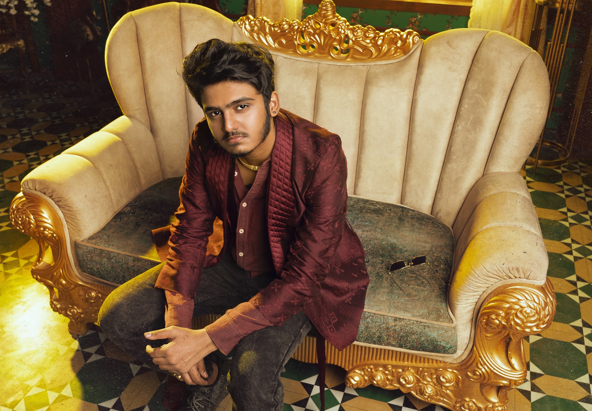 Sai Abhyankkar in red jacket sitting on a gold, white sofa for Katchi Sera video