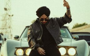 Singhsta sitting on a car in a black jacket, wearing sunglasses and raising one hand in the air