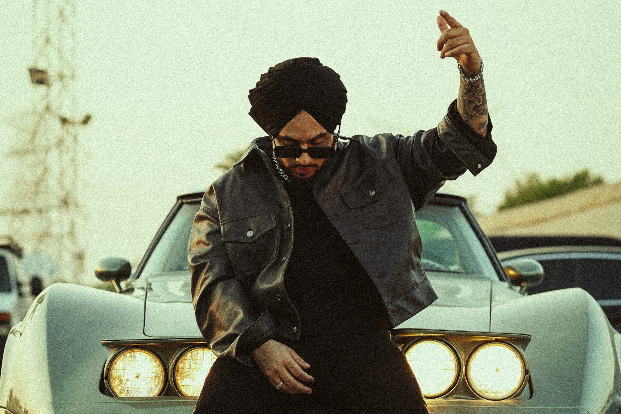 Singhsta sitting on a car in a black jacket, wearing sunglasses and raising one hand in the air