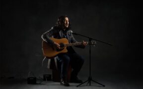 Sahitya Nepal Yaatri artist wearing black outfit, sitting and playing acoustic guitar