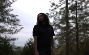 Rapper Bandzo3rd with long hair wearing black in a forest