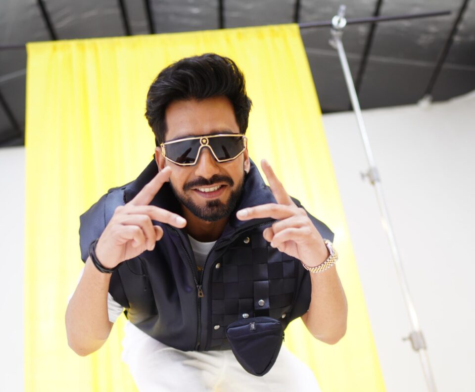 Maninder Buttar smiling and making hand signals wearing black sunglasses and black jacket with white pants