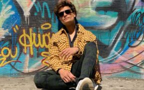 A-Zal seated on the ground wearing a yellow jacket, sunglassses and with graffiti on the wall behind him