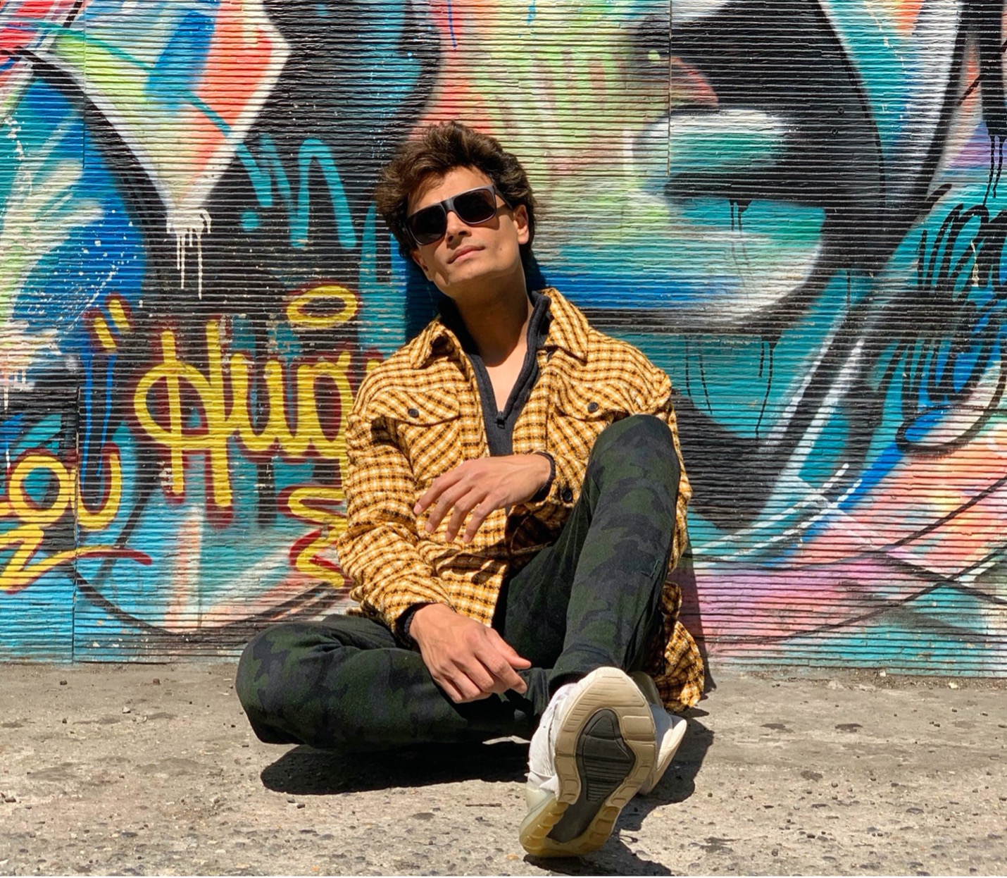 A-Zal seated on the ground wearing a yellow jacket, sunglassses and with graffiti on the wall behind him