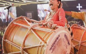 Purva Mantri smiling in red sari, beating dhol drum
