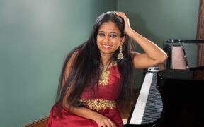 Charu Suri seated in a red dress with hand on piano