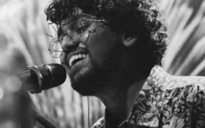 Dorwin John smiling, wearing glasses and singing into a mic