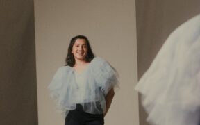 Frizzell D'Souza standing and looking into a mirror in white frilly top and black trousers