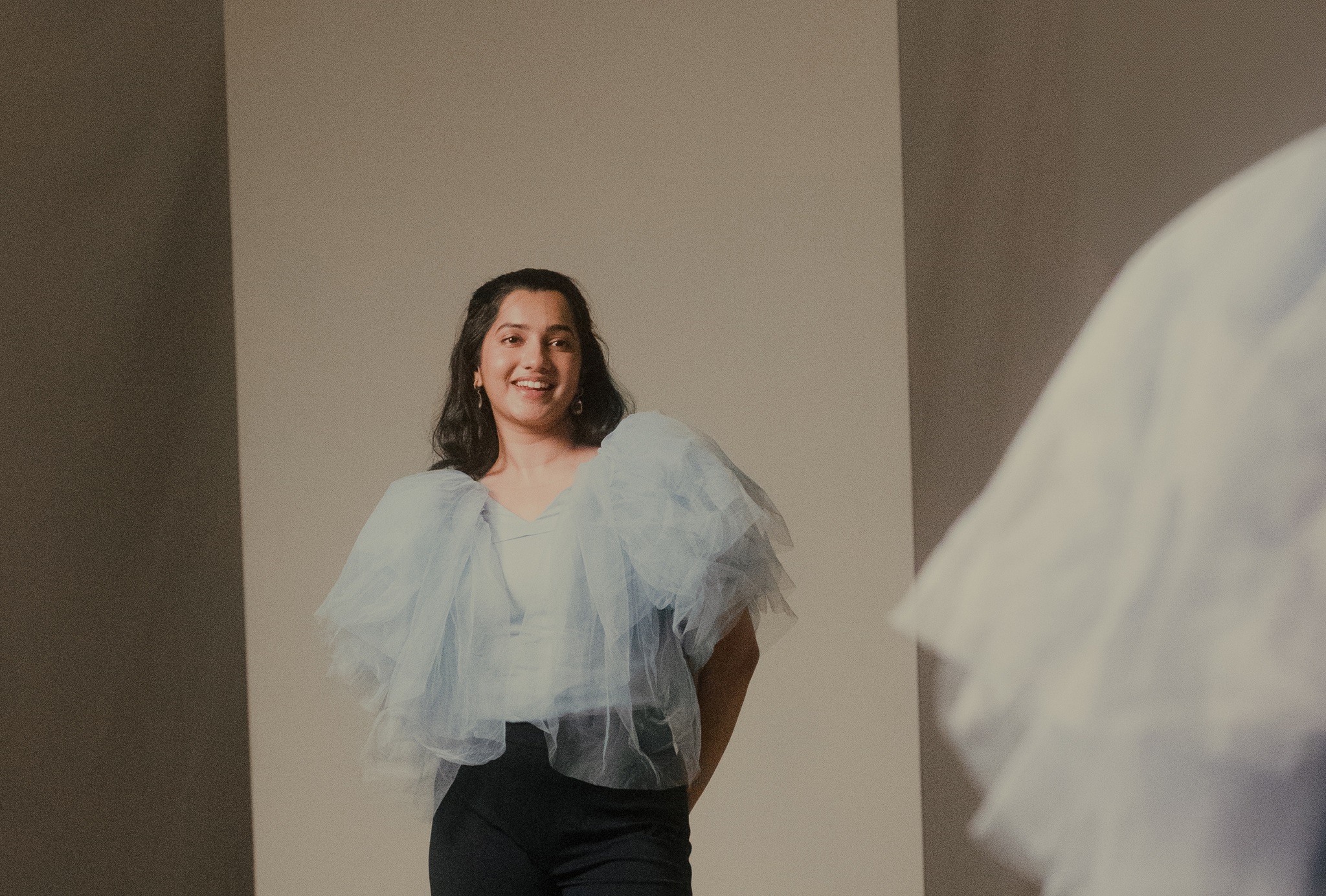 Frizzell D'Souza standing and looking into a mirror in white frilly top and black trousers