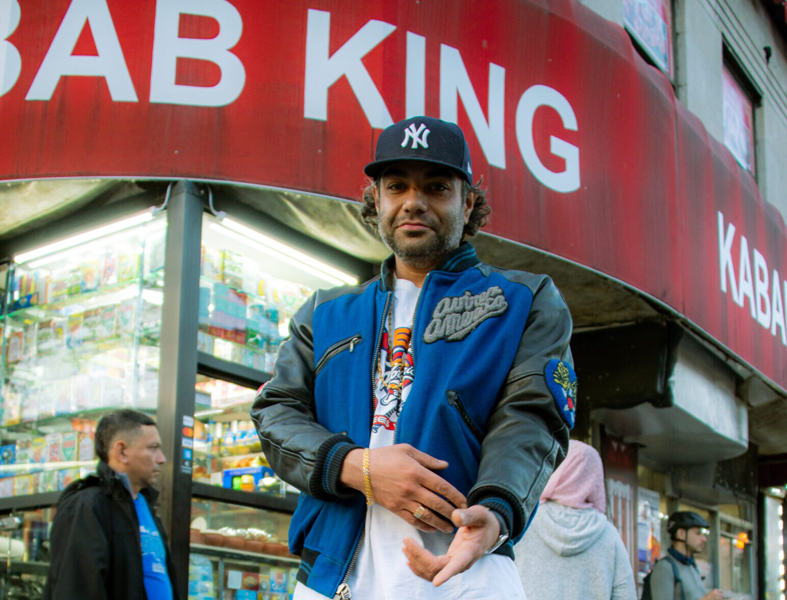 New York artist Heems aka Himanshu Suri in blue jacket and black cap standing outside a kebab shop