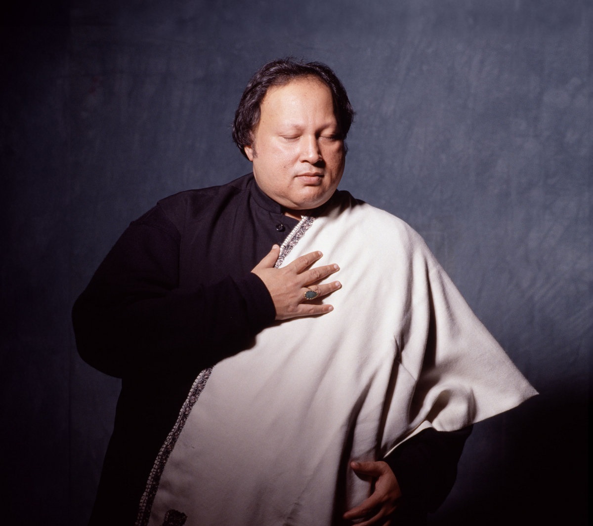 Nusrat Fateh Ali Khan in a black shirt with white shawl holding his hand to chest, with eyes closed