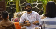 Prabh Deep wearing white and a grey hat, with glasses smiling and playing a ukulele