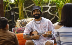 Prabh Deep wearing white and a grey hat, with glasses smiling and playing a ukulele