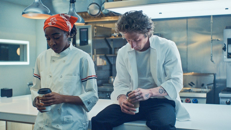 Syd and Carmy in their white chef outfits speaking in a kitchen area