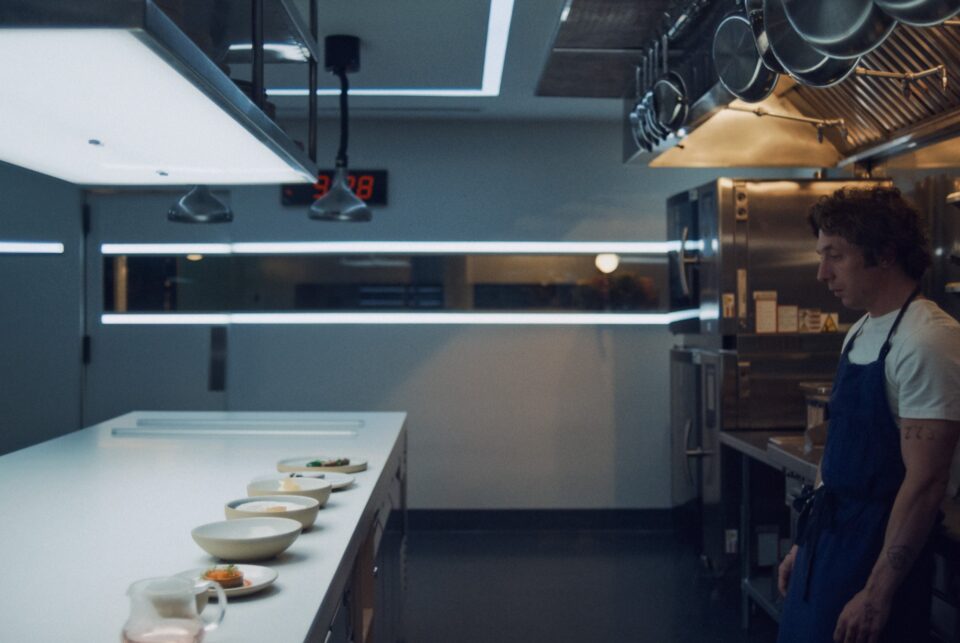 Carmy wearing a blue apron over a white tshirt approaches the kitchen counter under lights