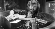 Kris Kristofferson seated on a couch playing guitar, with his shirt open