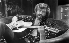 Kris Kristofferson seated on a couch playing guitar, with his shirt open