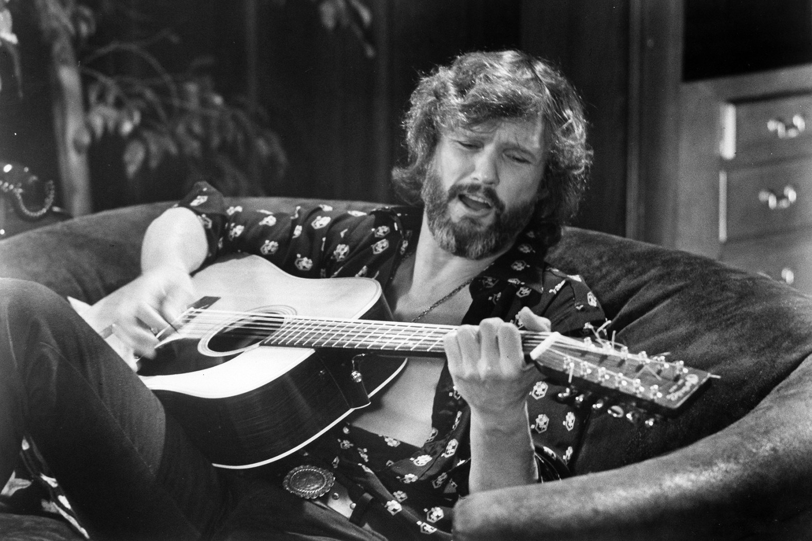 Kris Kristofferson seated on a couch playing guitar, with his shirt open