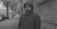 Shashwat Bulusu wearing black and a hat standing in front of a brick wall