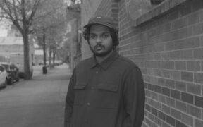 Shashwat Bulusu wearing black and a hat standing in front of a brick wall