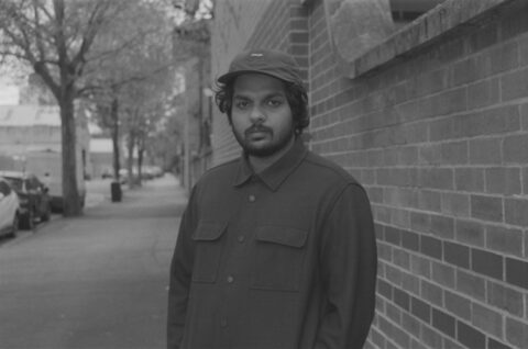 Shashwat Bulusu wearing black and a hat standing in front of a brick wall