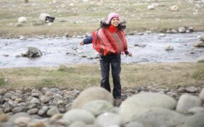 Amritha Rajan in a red multicolor sweater running among rocks near a water body