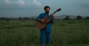 Singer-songwriter Bhrigu Sahni wearing a blue shirt and trousers standing in a green field playing guitar