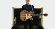 Bryan Adams standing in the trunk of a car with a brown guitar