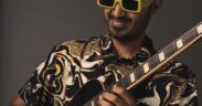 Chirag Todi in a black print shirt, wearing yellow rimmed glasses and playing guitar