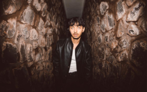 Raman wearing black jacket with white T-shirt surrounded by walls with rocks on them