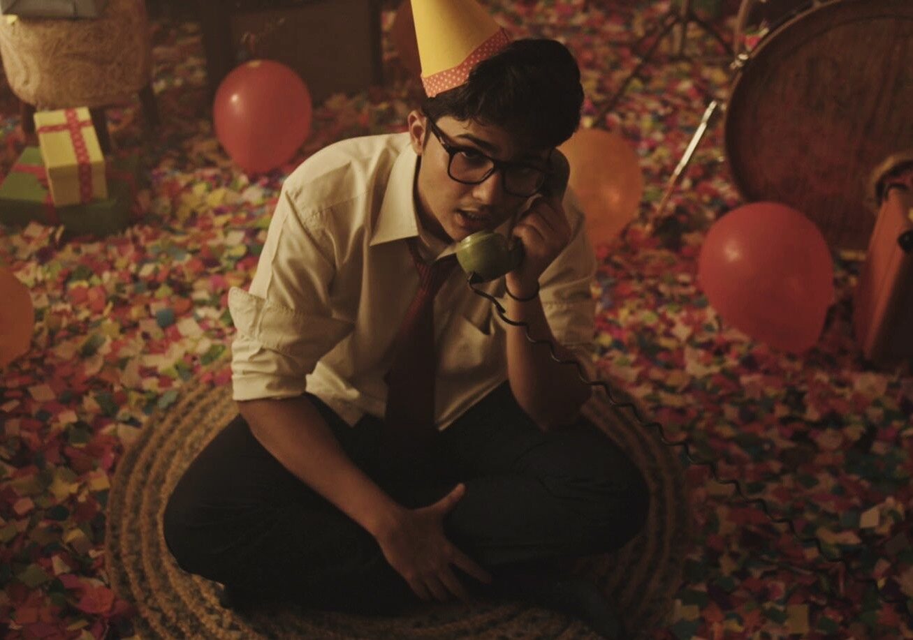 Janisht Joshi wearing a cone cap, white shirt, tie and black trousers, speaking into a telephone