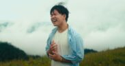 Markio Tanaldo in a blue shirt over a white t-shirt singing among green hills and clouds