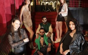 Indian rappers The Shiv in glasses and green jacket and Muhfaad seated on stairs with women surrounding them in club setting