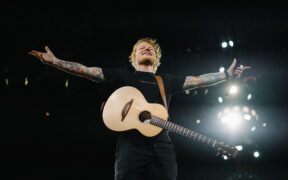 Ed Sheeran throwing his arms wide open on stage with a guitar