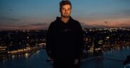 Martin Garrix wearing black standing with a city backdrop behind him