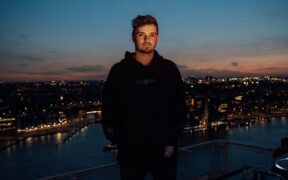 Martin Garrix wearing black standing with a city backdrop behind him