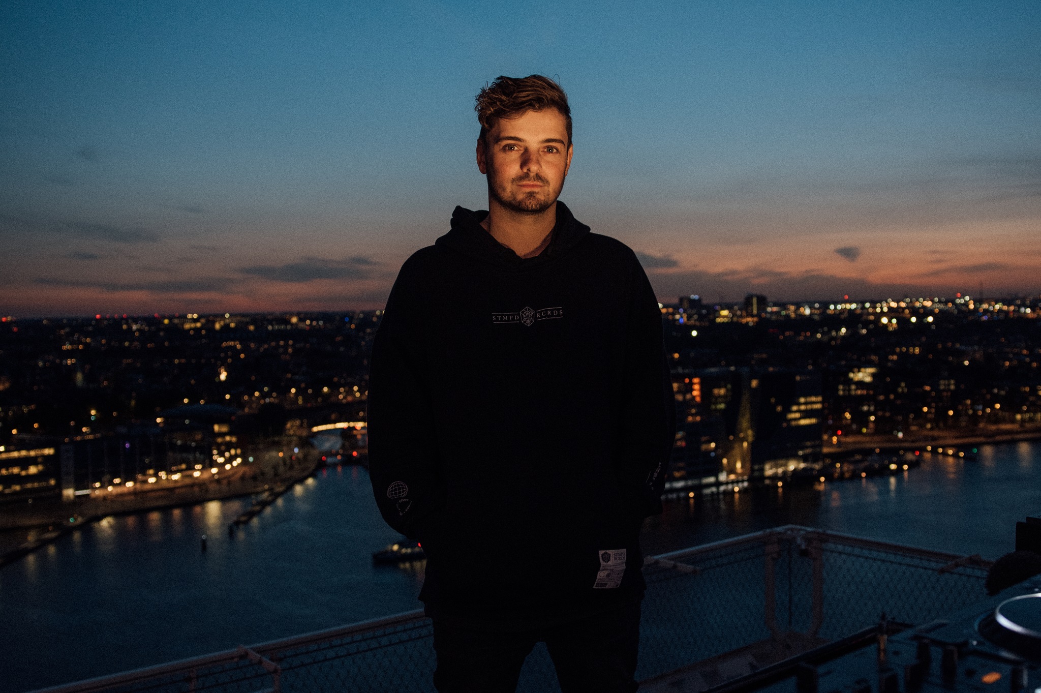 Martin Garrix wearing black standing with a city backdrop behind him