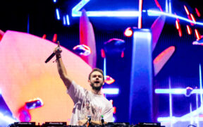 Zedd performing on stage with a DJ deck raising his hand with a mic