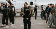 Parmish Verma wearing black t-shirt, trousers and brown jacket, walking on a tarmac with a private jet behind him