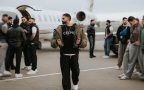 Parmish Verma wearing black t-shirt, trousers and brown jacket, walking on a tarmac with a private jet behind him