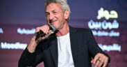 Sean Penn wearing white tshirt and black jacket and smiling as he speaks into a microphone, cigarette in hand